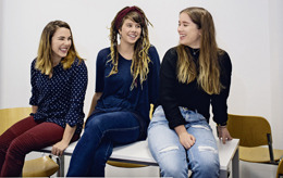 Drei Erasmus-Studentinnen aus Berlin sitzen nebeneinander auf einem Tisch in einem Seminarraum ihrer Hochschule und lachen sich an.